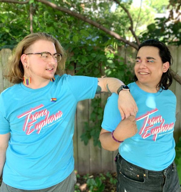 Two trans people, one white with long blond hair and rainbow dangle earrings, and one Latinx with light brown skin, long dark brown hair and trans-flag-colored eye shadow. They look at each other, smiling and holding hands. They both wear bright blue t-shirts that say "Trans Euphoria."