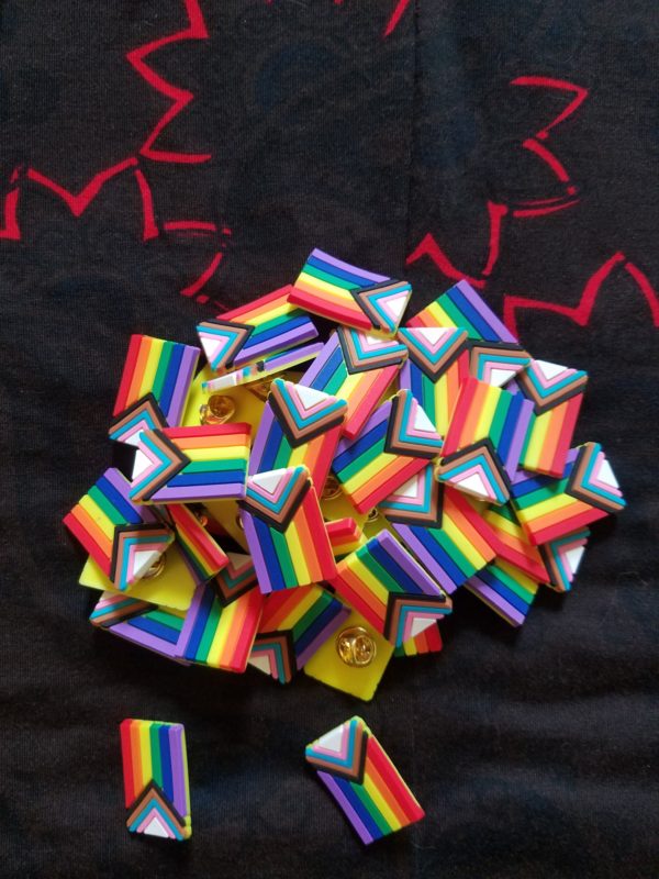 A pile of progress pride flag lapel pins on a black background.