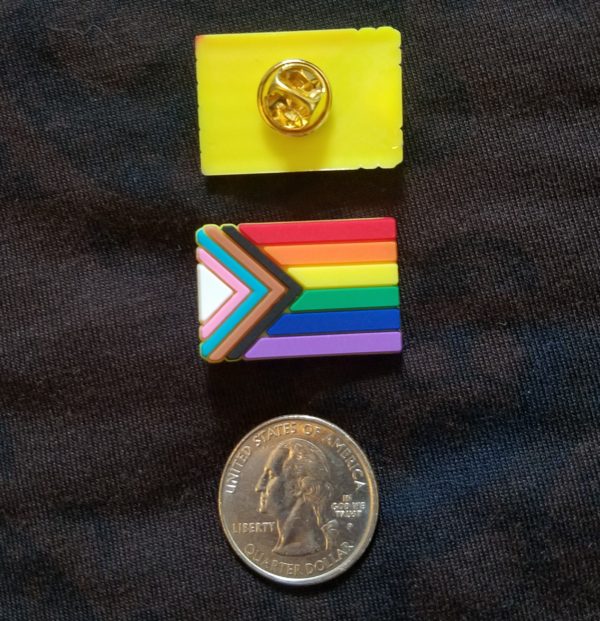 A progress pride flag lapel pin next to a quarter for size comparison. They are roughly the same size.