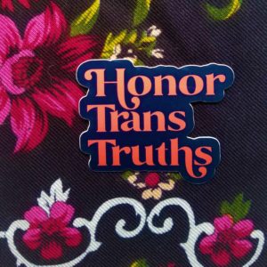 Set against a rich dark floral cloth, sits a dark blue vinyl sticker that say Honor Trans Truths in an deep coral embellished font.