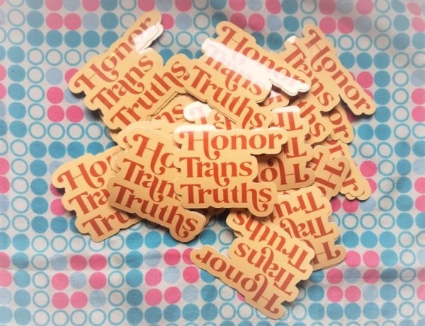 Set against a bubbly pink white and blue fabric, sits a stack of cream colored vinyl stickers that say Honor Trans Truths in an burnt orange embellished font.