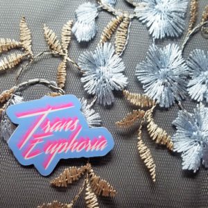 Set against a black mesh fabric with shimmery gold and silver embroidered flowers, sits a light blue vinyl sticker that says Trans Euphoria in bright white scrawling text with a bright pink shadow.