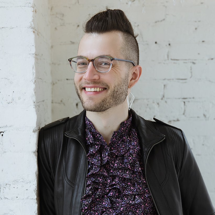 Photo of Ximón leaning against a white brick wall. They are a tall light-skinned person with glasses, a short-trimmed beard, and have a small bouffant with shaved sides, wearing a purple floral ruffled shirt with a black leather jacket over top. They smile widely..