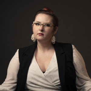 A photograph of Nicholette Driggs (she/her). She wears a flowing long-sleeve v-neck white blouse with shimmery gold speckles, topped with a simple black vest. Her long brown hair is pulled up into a flat bun. She wears shimmery cat-eye glasses, and has two dangle earrings that consists of multiple loops of pearls.