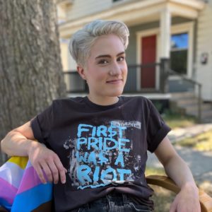 Photo of Arlo Van Horn. They are a light-skinned person, young, with swooped blond hair with a buzzed side. They smile pleasantly. They are wearing a black t-shirt that says First Pride was a Riot.