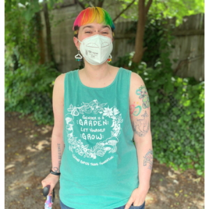 Photo of a masked model with rainbow hair and a cane in a seafoam tanktop.