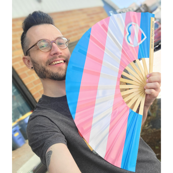 Photo of Ximon holding a clack fan in trans flag colors