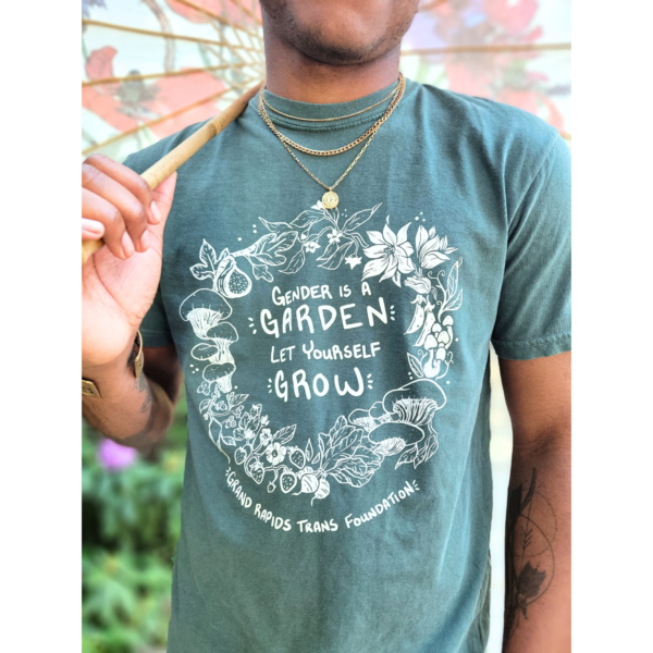 A close-up photo a dark bluish-green t-shirt that has white text that reads "Gender is a Garden, Let Yourself Grow" surrounded by a wreath of botanicals, including flowers, mushrooms, radishes, strawberries, and peas. Under the wreath design reads Grand Rapids Trans Foundation. The model, Artemis, is holding a floral umbrella.