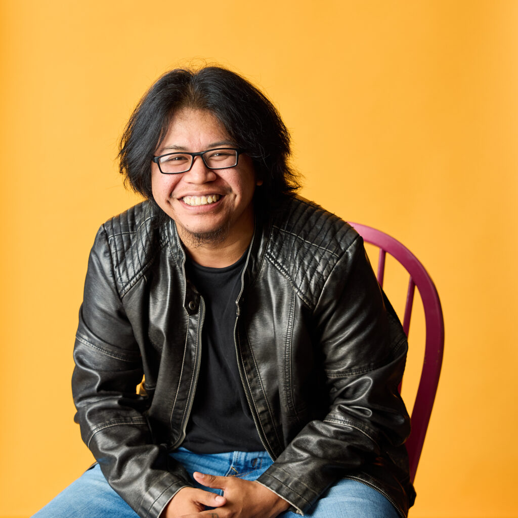 Photo of Alexander, seated. He is a caramel-skinned person with a round face and dimples, with shoulder-length straight hair, wearing a black shirt and a black jacket. He smiles widely.