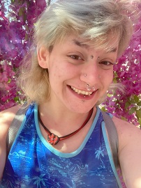 A picture of Hayden Ryan (he/they), a white Trans-masculine person with a light blonde mullet hairstyle, brown eyes, and multiple facial piercings. Hayden is smiling wide against a background of dark pink flowers and is wearing a blue tank top with a palm tree print.