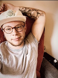 A picture of Kasey Powlenko (he/him), a white Trans man wearing tortoise shell glasses, a cream colored hat reading "Outside Coffee Co." and a grey pocket tee-shirt. He is smiling pleasantly with one arm featuring a tattoo of a tree resting behind his head.
