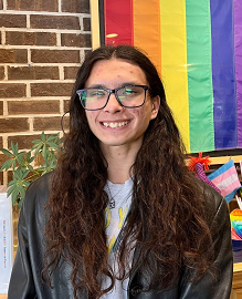 A picture of Lex Larkspur (she/her), an Asian Trans woman with dark brown long curly hair wearing dark rectangular glasses and a grey t-shirt with a black leather jacket. She is smiling pleasantly against a brick wall with a rainbow flag hanging.