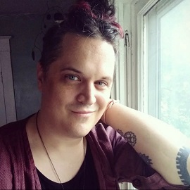 A picture of Rickey Ainsworth (they/them), a white Non-binary person wearing a dark magenta cardigan over a black top. They have dark hair with magenta accents pulled into a high bun and they are smiling with one hand resting on the side of their face.